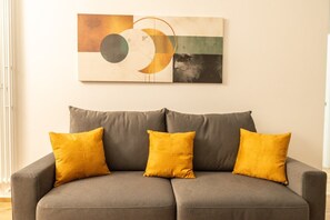 Un canapé gris agrémenté de trois coussins de couleur jaune moutarde, un tableau abstrait accroché au mur et une fenêtre avec des stores.
