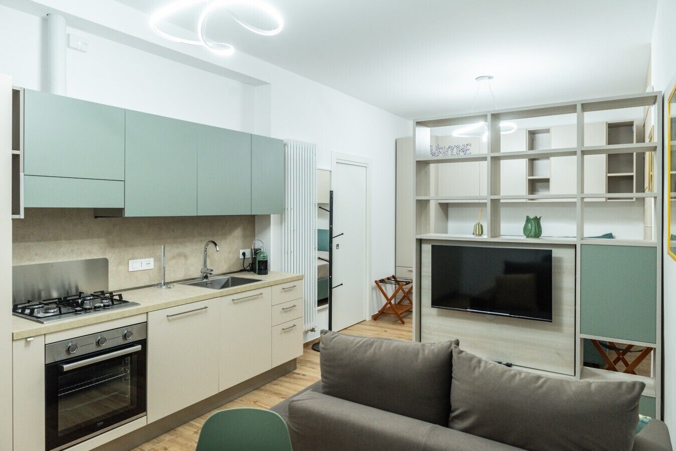 A modern kitchen with a built-in oven, a gas stove, and a sink. There is a flat-screen TV mounted on a wall, and a grey sofa in the living area.
