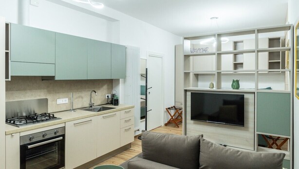 Een moderne keuken met een ingebouwde oven, een gasfornuis en een spoelbak. Er is een flatscreen televisie aan de muur bevestigd en een grijze bank in de woonkamer.