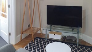 A living room with a TV on a glass stand, a wooden hanger shelf, a patterned rug, and a white table. - Kinamara Ensuite Room (Margate)