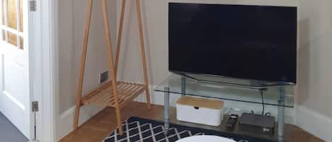 Una sala de estar con un televisor sobre un pedestal de vidrio, un estante de madera para perchas, una alfombra estampada y una mesa blanca.