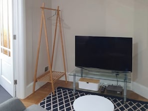 Uma sala de estar com uma TV sobre um suporte de vidro, uma estante de cabides de madeira, um tapete estampado e uma mesa branca.