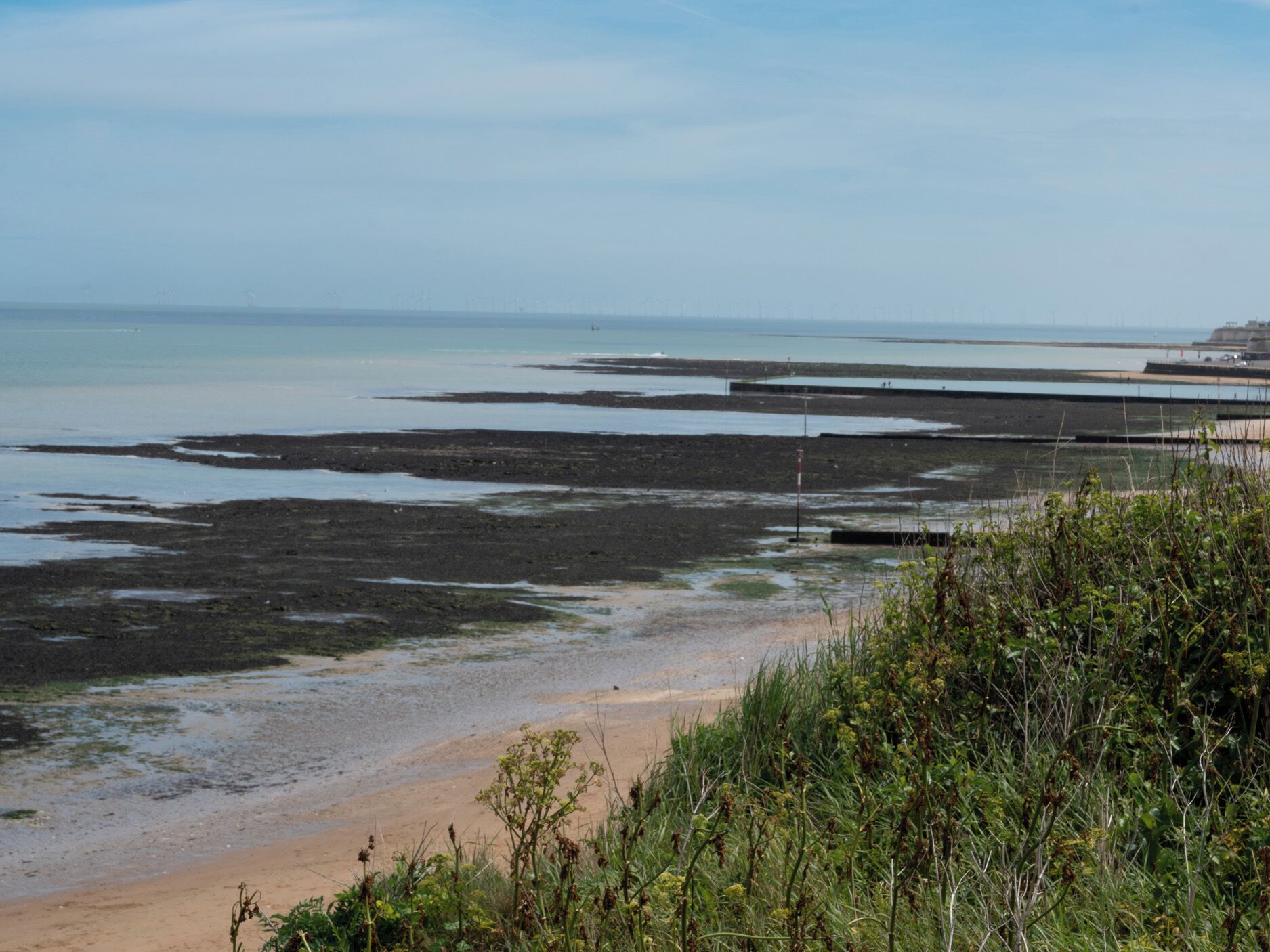 Un paisaje costero con una playa arenosa, un litoral rocoso y una vista distante de un muelle.