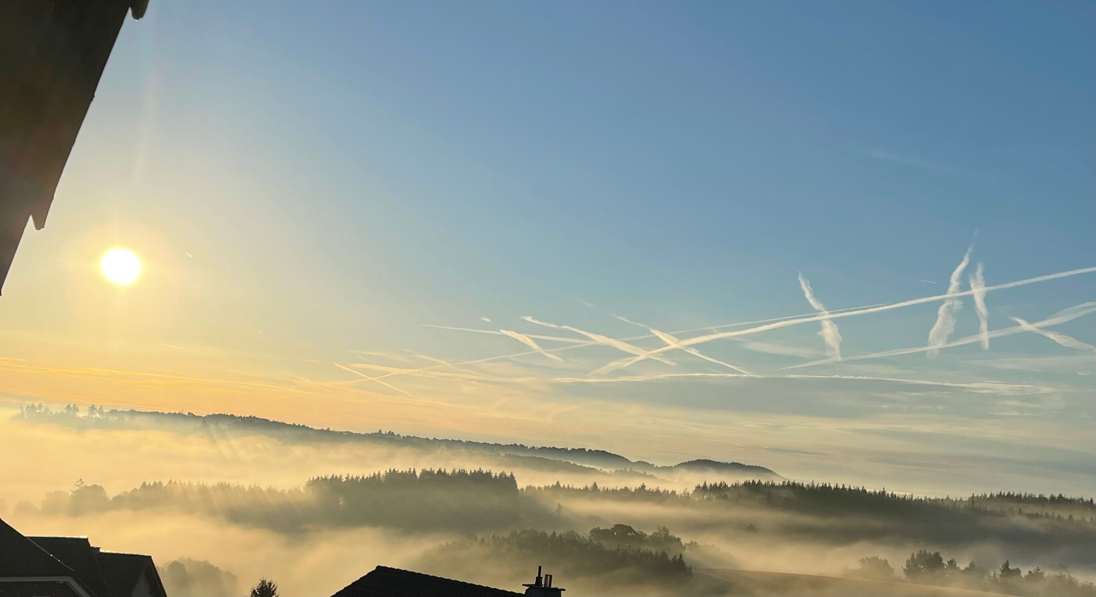 A sunrise over a misty landscape with visible contrails in the sky.