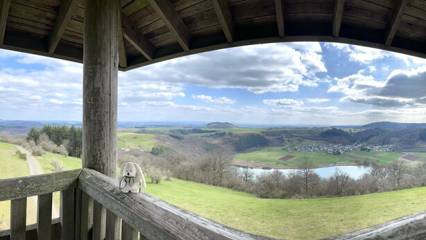 A wooden balcony with a view of a rural landscape, including fields, a lake, and a small town.