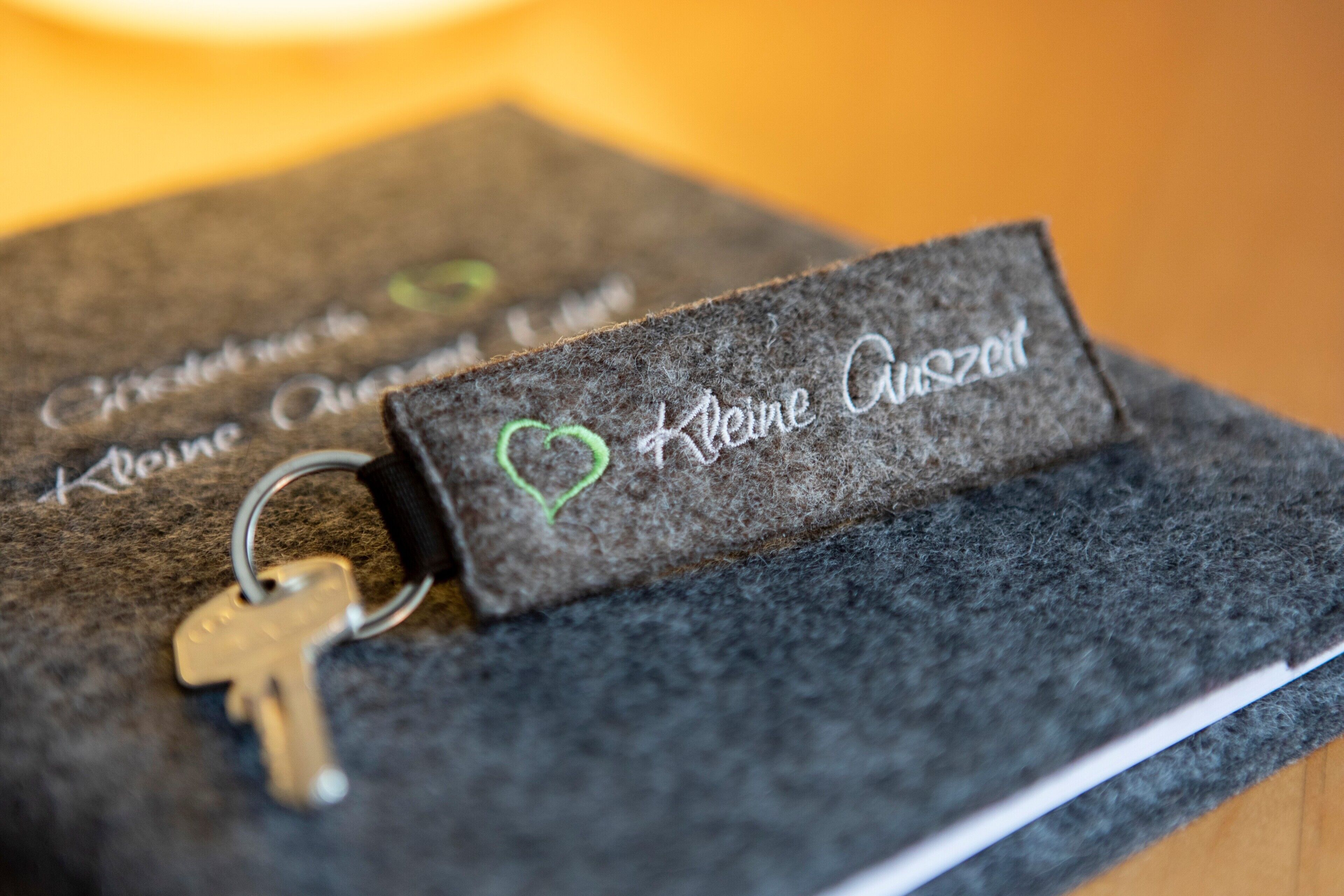 Un llavero con un sujetapersonas en forma de corazón y una llave, sobre una superficie oscura y texturizada. Se lee el texto 'Kleine Auzeit'.