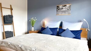 A neatly made bed with a white and gray patterned blanket, a wooden ladder, a small table with a vase of flowers, and a wall-mounted picture frame.