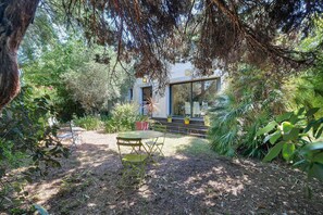 Um jardim com mesa e cadeiras, um prédio com porta de vidro e uma árvore ao primeiro plano.