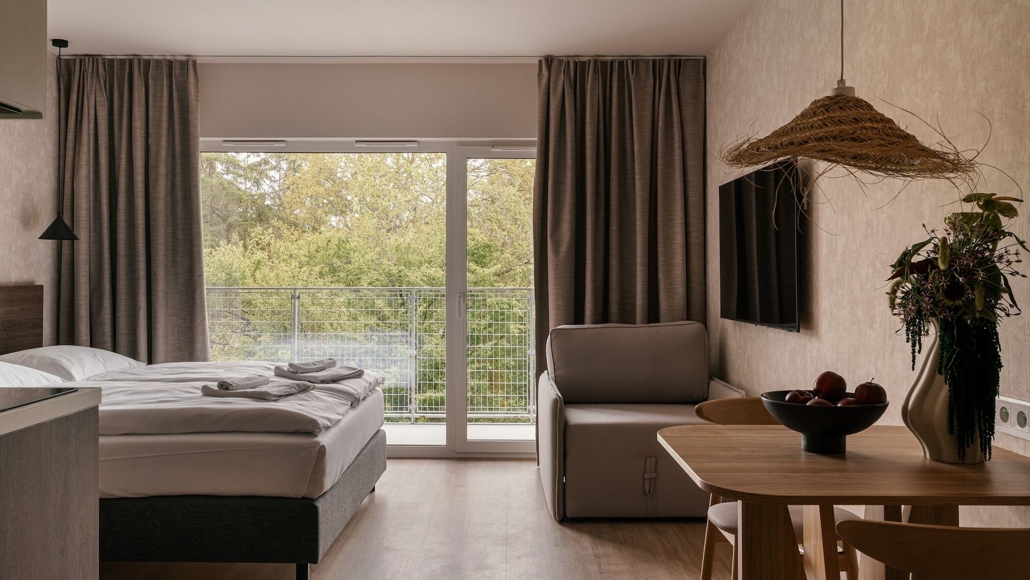 A modern hotel room with a bed, a sofa, a dining table, a plant, and a balcony with a view of trees.