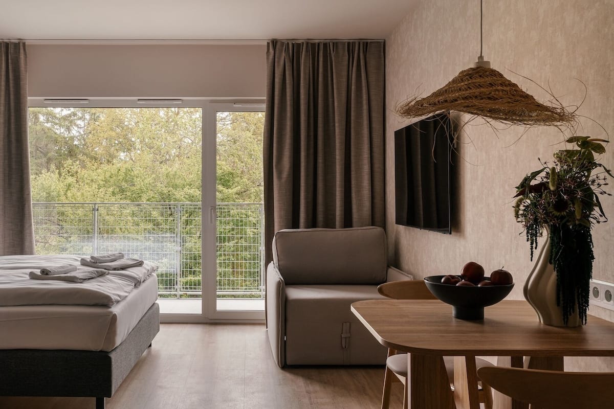 Ein modernes Hotelzimmer mit Bett, Sofa, Esstisch, einer Pflanze und einem Balkon mit Blick auf Bäume.