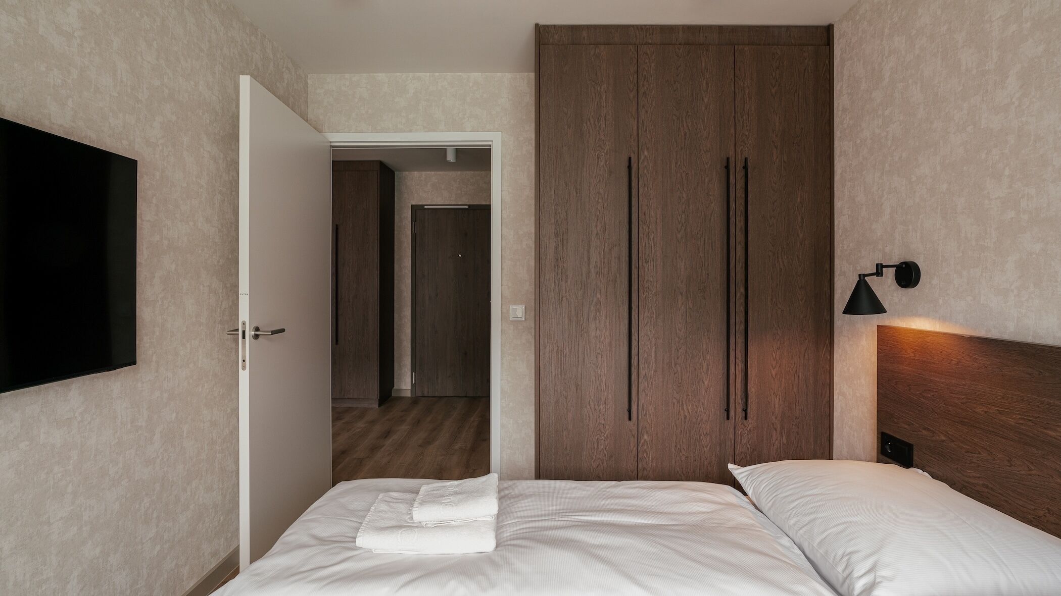 A hotel room with a bed, a TV, a wardrobe, and a wall-mounted lamp.