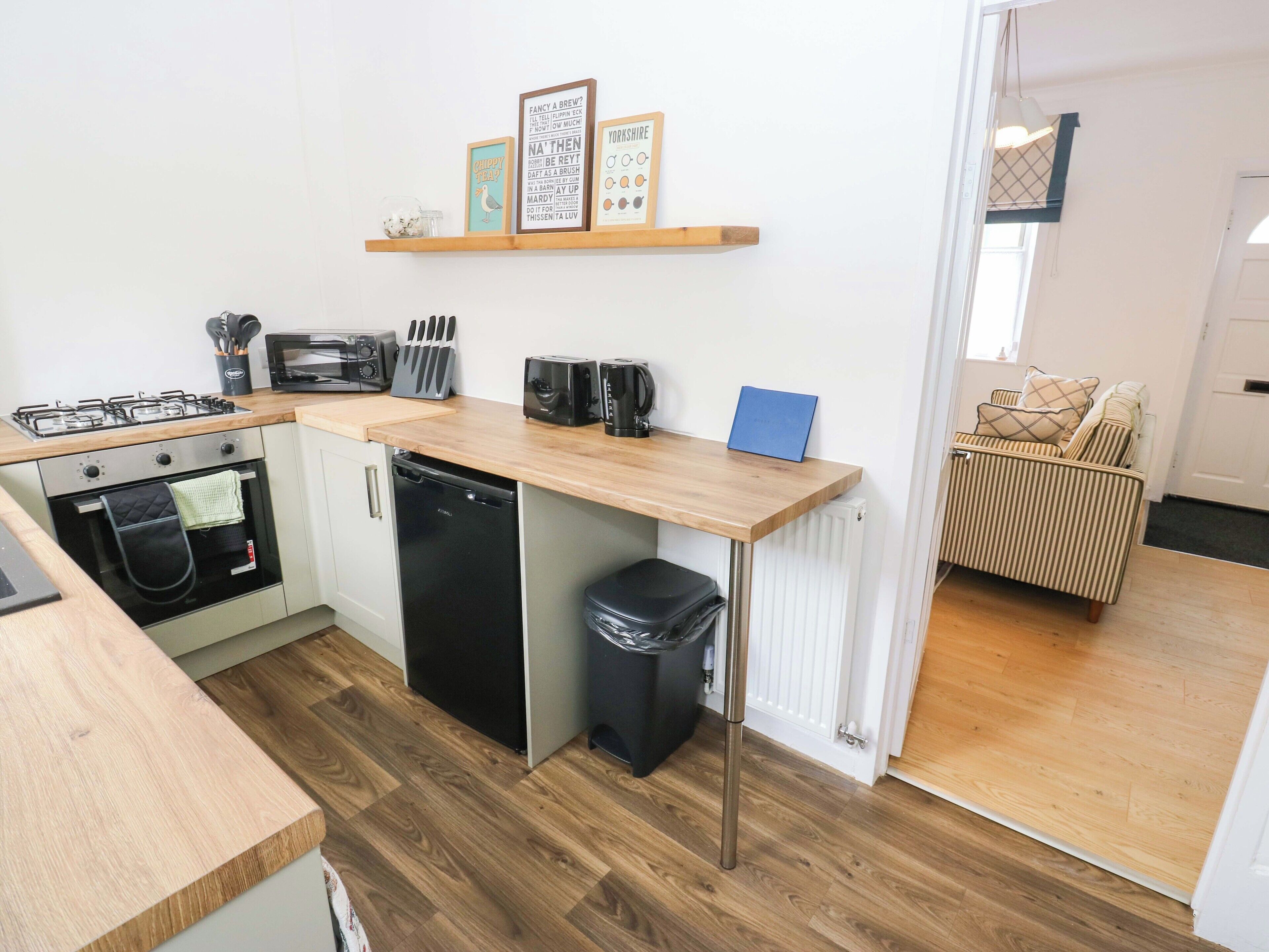 Una cocina funcional equipada con estufa a gas, horno, lavavajillas y cafetera. Se observa un cubo de basura pequeño y una repisa con objetos decorativos.