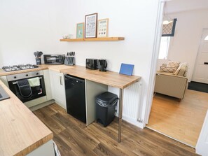 A compact kitchen with a gas stove, oven, dishwasher, and a coffee maker. There is a small trash bin and a shelf with decorative items. - Saltaire House (Shipley)