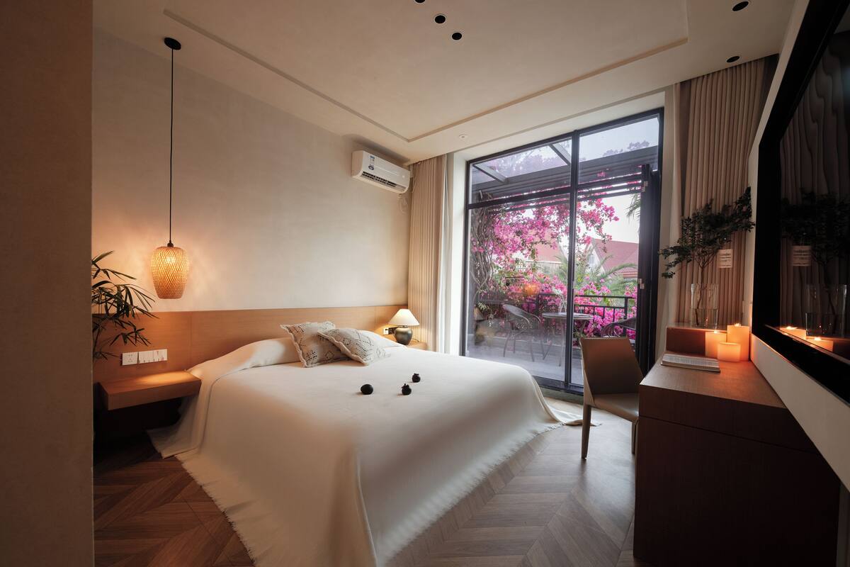 Et moderne hotelværelse med en stor seng, et skrivebord, en stol, et vindue med udsigt til lyserøde blomster og en hængende lampe.