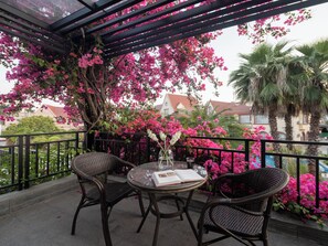 A covered outdoor seating area with wicker chairs and a table, surrounded by pink bougainvillea flowers and palm trees.