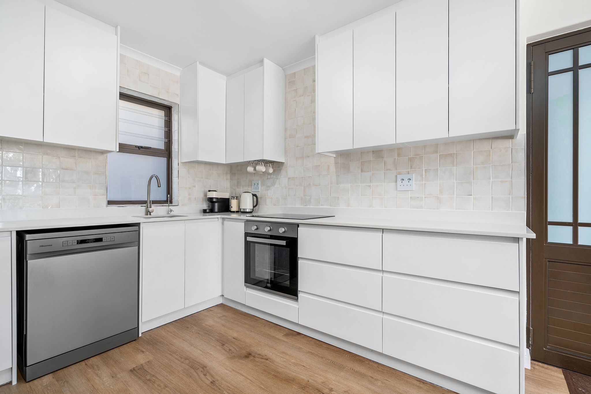 Eine moderne Küche mit weißen Schränken, einem Geschirrspüler, einem Backofen und einem Fenster mit Jalousien.