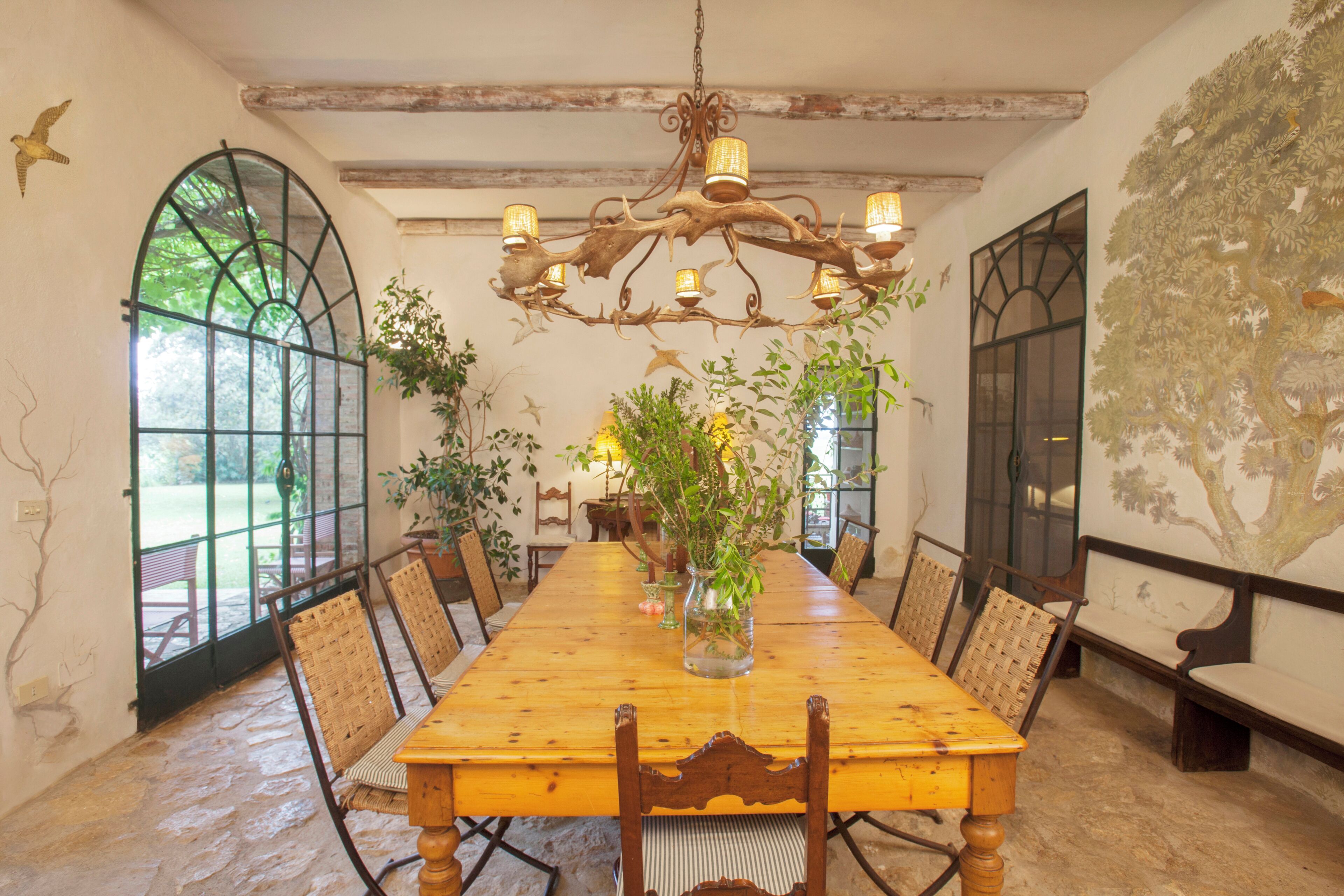 A dining room with a long wooden table, chairs, a chandelier, and large windows.