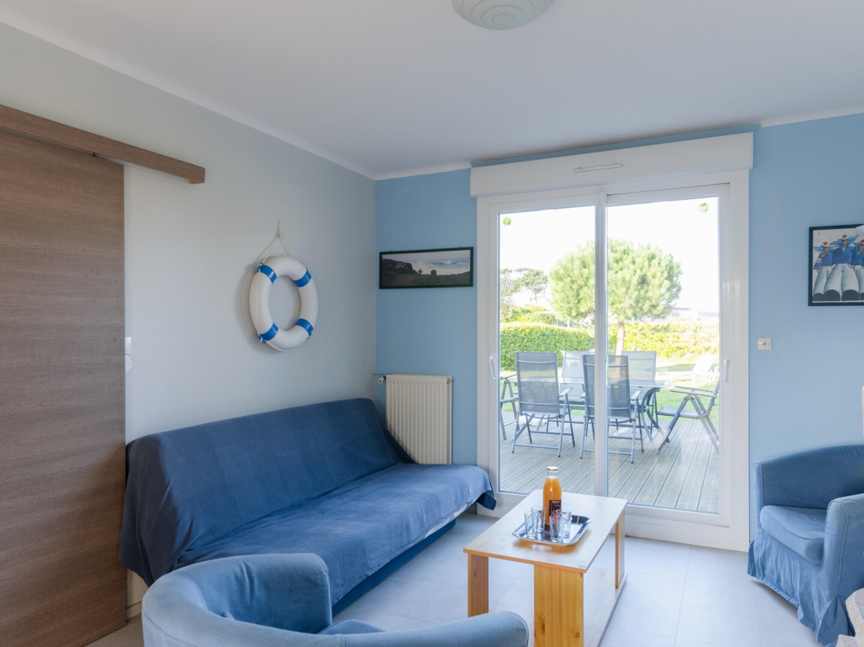 A room with a blue sofa, a small table with a tray, a life ring, and a view through a glass door to an outdoor area with chairs.