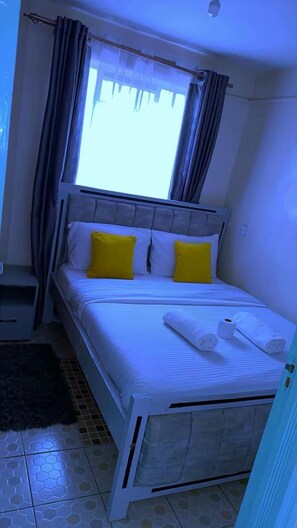 A single bed with white bedding and two yellow pillows, a bedside table, and a window with curtains. - Cozy 1BR Home Away From Home with Balcony Near TRM Mall and Garden City (Nairobi)