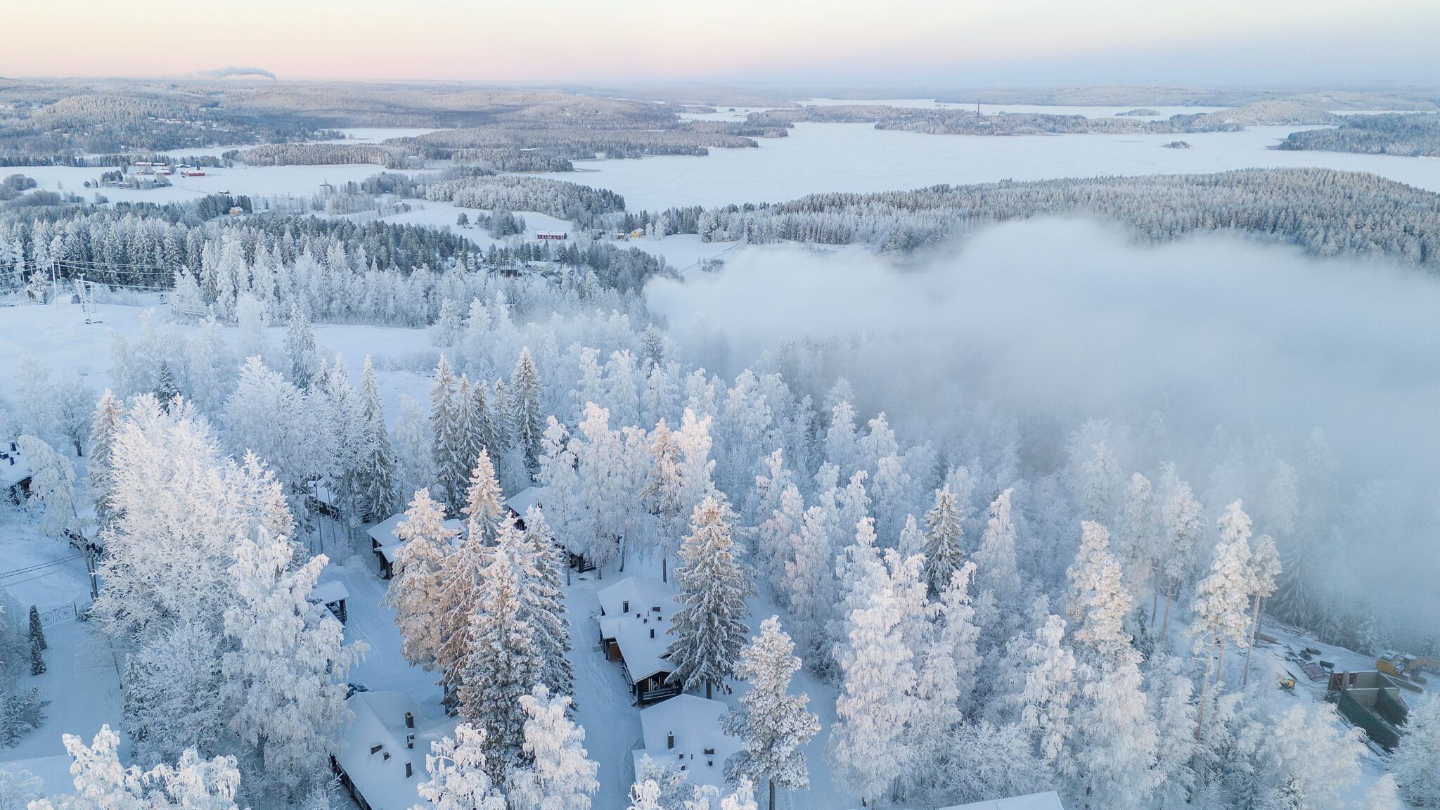 Un bosque cubierto de nieve con cabañas, un lago y un fondo brumoso.