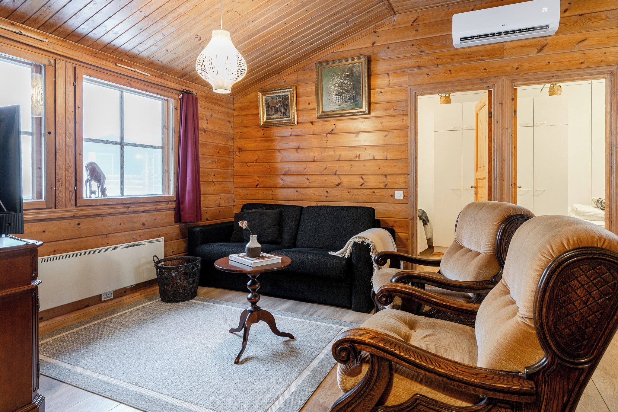 Una habitación con paneles de madera, un sofá negro, dos sillones de madera, una pequeña mesa con un florero y una lámpara colgante.