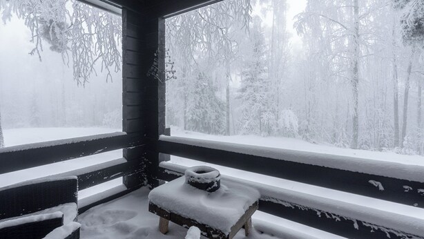 Un banco de madera cubierto de nieve frente a una ventana con vistas a un bosque nevado.