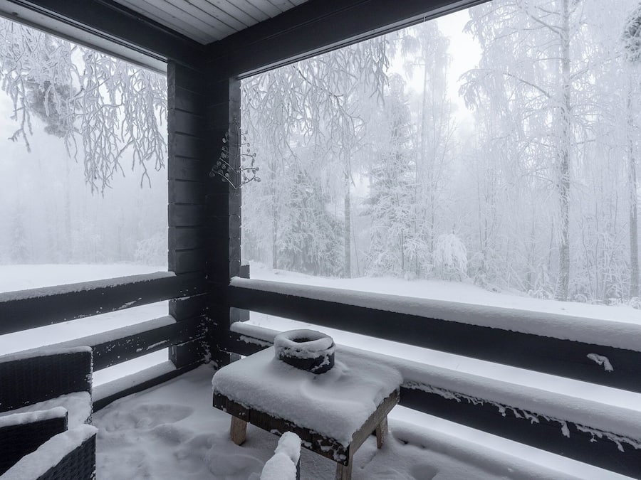 Un banco de madera cubierto de nieve frente a una ventana con vistas a un bosque nevado.