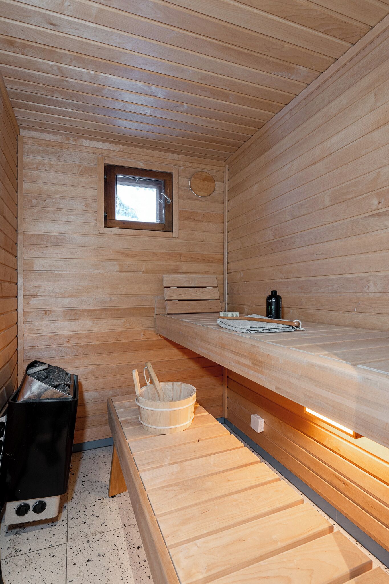 A wooden sauna room with a bench, a small window, a heater, and a bucket.