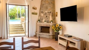 A dining area with a wooden table and chairs, a stone fireplace, a flat-screen TV, and a view of a pool through a glass door. - Velletri, elegant guesthouse with pool (Greta's Villa) (Velletri)