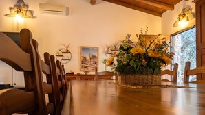 A dining area with wooden chairs, a central table with a woven basket of flowers, wall-mounted air conditioning, and a framed desert landscape painting. - Velletri, elegant guesthouse with pool (Greta's Villa) (Velletri)