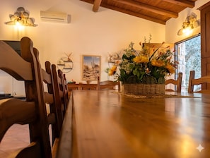 A dining area with wooden chairs, a central table with a woven basket of flowers, wall-mounted air conditioning, and a framed desert landscape painting.