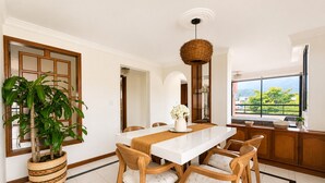 Un coin repas avec une table blanche, des chaises en bois, un luminaire suspendu en fibres tressées et une vue sur la verdure à l’extérieur.