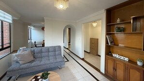 Un salon avec un canapé gris, une table basse en bois, un lustre et un meuble en bois sur lequel est posé un panneau indiquant « Bienvenue ».