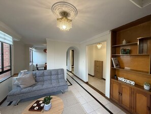 Ein Wohnzimmer mit grauem Sofa, einem Holztisch, einer Kristallleuchtre und einer Holzkassette mit der Aufschrift 'welcome'.