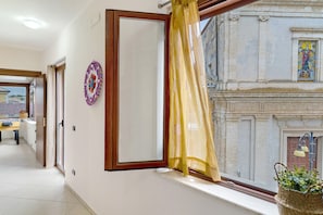 A room with a window, a mirror, a curtain, and a view of a building with a statue. - La Chiazza di Grutti – Historic Center, Wi-Fi & Air Conditioning (Grotte)