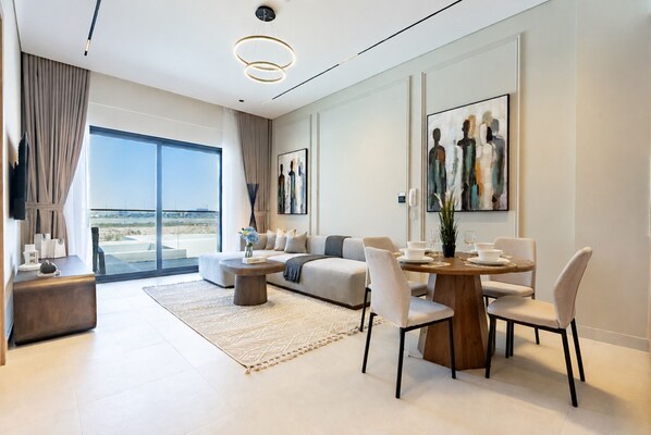 A modern living room with a sofa, dining table, chairs, and artwork. - Serene 1BR with Assistant Room, Only Minutes by Car from DAMAC Hills Mall (Dubai)