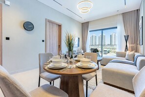 A modern hotel room with a dining area, a sofa, a large window with a city view, and a clock on the wall. - Serene 1BR with Assistant Room, Only Minutes by Car from DAMAC Hills Mall (Dubai)