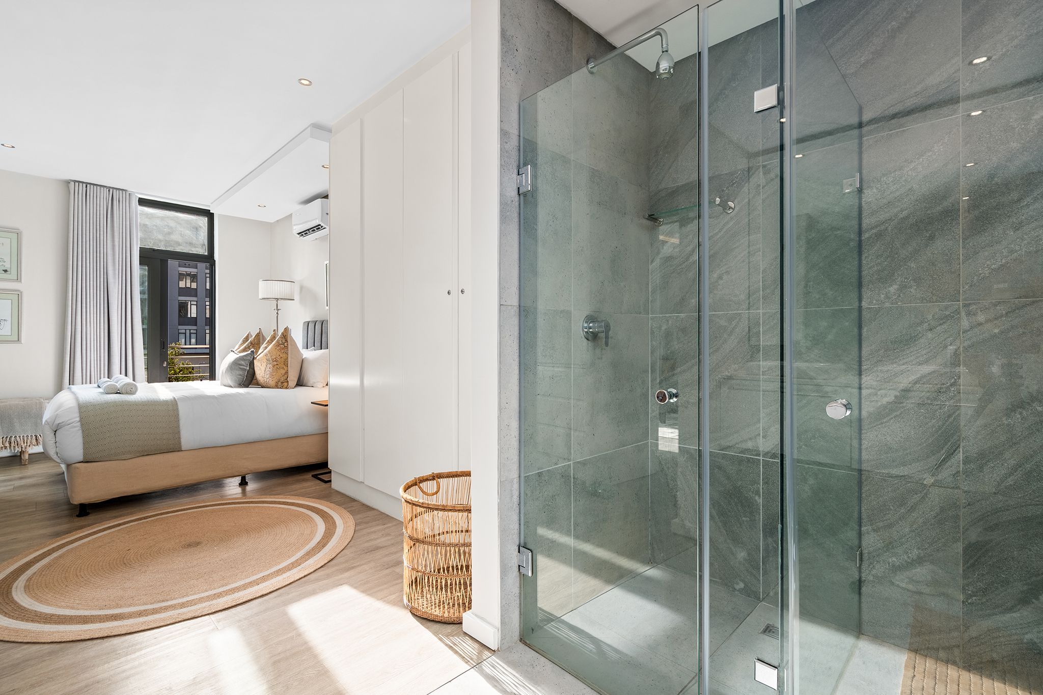 A modern bedroom with a glass-enclosed shower, a bed with a view, and a wicker basket.
