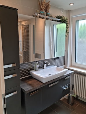 Ein modernes Badezimmer mit großem Spiegel, weißem Waschbecken, dunklen Schränken und einem Fenster für natürliches Licht.