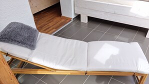 A room with a wooden-framed lounge chair, a grey fluffy pillow, and a white bench against a white wall.