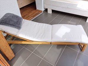 A room with a wooden-framed lounge chair, a grey fluffy pillow, and a white bench against a white wall.