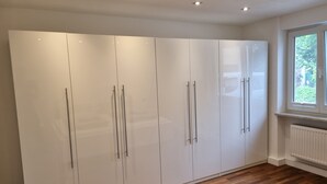 A room with a large white wardrobe, wooden flooring, and a window with a view of greenery.