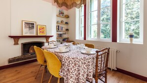 Una sala menjador amb una taula posta per a quatre, una llar de foc, prestatges de llibres i un gran finestral amb vista a arbres.