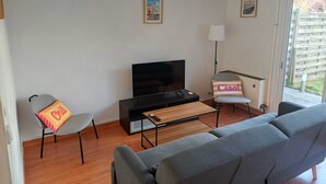 A living room with a sofa, a TV, a coffee table, and a chair with a cushion. There are two framed pictures on the wall.