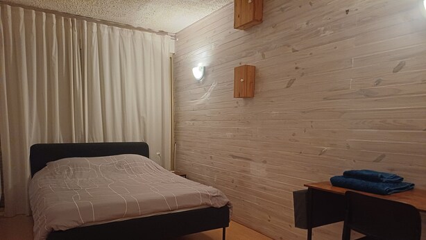 A bedroom with a bed, a desk, a chair, a wooden wall, and a window with curtains.