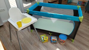 A baby playpen with a high chair beside it, a puzzle box, and a toy on the floor. - Apartment Abel with Balcony, Wi-Fi, Charging Station & AC near Hospital and Rehab Center (Oldenburg)