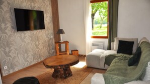 A living room with a flat-screen TV, a round wooden coffee table, a sofa, a lamp, and a view of greenery outside. - O Reflection of the Lake (Groléjac)
