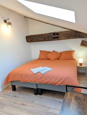 A bedroom with a bed, a skylight, a wooden beam, a nightstand with a lamp, and a small table. - Gîte du Bosquet (Oradour-sur-Glane)