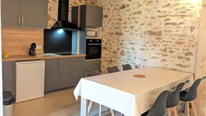 Een keuken met een stenen muur, houten balken in het plafond, grijze kastjes, een witte tafel en zwarte stoelen.