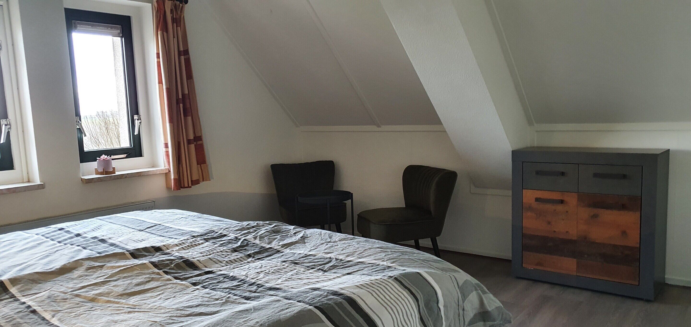 A bedroom with a bed, two chairs, a dresser, and a window with curtains.